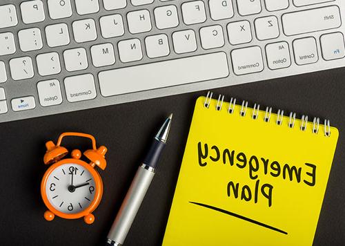 Emergency Plan writing on a yellow notepad placed next to a pen, alarm clock & keyboard